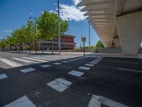 a car sitting at an empty street underneath a overpass overhang that contains a train stop, and cars and a pedestrian