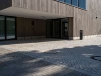 a building is shown with the entrance door open on the cobblestone path, with a trashcan on the sidewalk in front of it