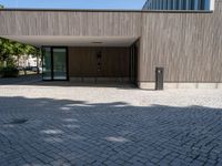 a stone patio with a bench outside of a large wooden building with windows that open onto a paved courtyard