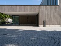 a stone patio with a bench outside of a large wooden building with windows that open onto a paved courtyard