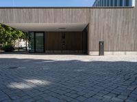 a stone patio with a bench outside of a large wooden building with windows that open onto a paved courtyard