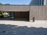 a stone patio with a bench outside of a large wooden building with windows that open onto a paved courtyard