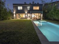 a swimming pool at night, near the house with lights on and patio furniture behind it