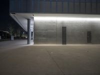 an outdoor walkway with lighting on it's sides at night with concrete walls and black metal doors