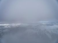 view from an airplane flying over a snowy mountains range in the foggy sky of the ocean