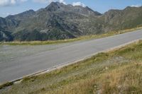 a motorcycle on a country road in front of mountains near grass and bushes, in front of a large mountain