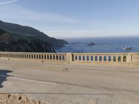 a man riding a motorcycle next to a long bridge over water with cliffs in the distance