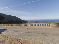 a man riding a motorcycle next to a long bridge over water with cliffs in the distance