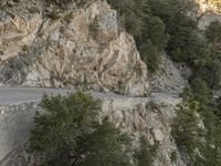 a person riding a motorcycle on a rock road next to trees and mountains with cliff cliffs