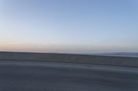 a person on a motorcycle rides along the side of a bridge at dusk, with the sun in the background