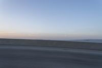 a person on a motorcycle rides along the side of a bridge at dusk, with the sun in the background