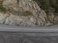 a motorcycle is driving down the mountain road near a cliff wall with trees around it