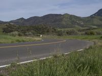 a motorcycle is parked on a rural road in the mountains near grass and trees with hills and bushes behind it