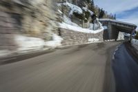 the speeding shot of a man driving his motorcycle down a mountain road with a bridge above