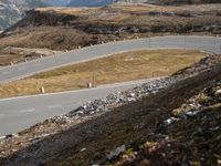 a motorcycle travels down the side of a mountain road in front of a fenced hill