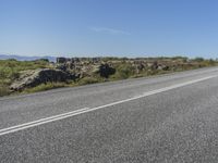 a motorcycle traveling down a highway beside grass and a cliff side area with rocks and shrubs