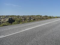 a motorcycle traveling down a highway beside grass and a cliff side area with rocks and shrubs