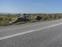 a motorcycle traveling down a highway beside grass and a cliff side area with rocks and shrubs