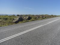 a motorcycle traveling down a highway beside grass and a cliff side area with rocks and shrubs