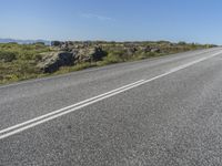 a motorcycle traveling down a highway beside grass and a cliff side area with rocks and shrubs