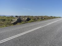 a motorcycle traveling down a highway beside grass and a cliff side area with rocks and shrubs