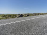 a motorcycle traveling down a highway beside grass and a cliff side area with rocks and shrubs