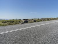 a motorcycle traveling down a highway beside grass and a cliff side area with rocks and shrubs