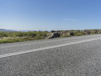 a motorcycle traveling down a highway beside grass and a cliff side area with rocks and shrubs