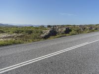 a motorcycle traveling down a highway beside grass and a cliff side area with rocks and shrubs