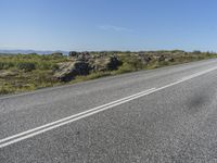 a motorcycle traveling down a highway beside grass and a cliff side area with rocks and shrubs