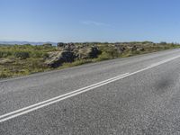 a motorcycle traveling down a highway beside grass and a cliff side area with rocks and shrubs