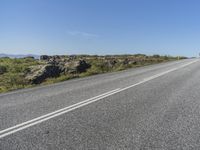 a motorcycle traveling down a highway beside grass and a cliff side area with rocks and shrubs