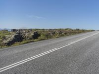 a motorcycle traveling down a highway beside grass and a cliff side area with rocks and shrubs