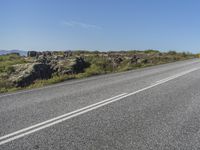 a motorcycle traveling down a highway beside grass and a cliff side area with rocks and shrubs