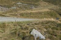 two motorcycles are parked on the side of a road in front of a hill, with grass and rocks scattered around the road