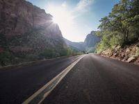 an empty road on a mountain side with trees on both sides of the road and a sun shine behind