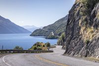 the man is traveling his motorcycle along a winding road beside the water and mountains on the other side