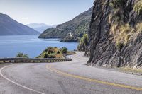 the man is traveling his motorcycle along a winding road beside the water and mountains on the other side