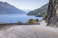 the man is traveling his motorcycle along a winding road beside the water and mountains on the other side