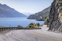 the man is traveling his motorcycle along a winding road beside the water and mountains on the other side