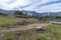 a dirt path going across a grassy valley to a mountain range with pine trees and snow capped mountain tops