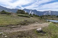 a dirt path going across a grassy valley to a mountain range with pine trees and snow capped mountain tops