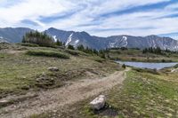 a dirt path going across a grassy valley to a mountain range with pine trees and snow capped mountain tops