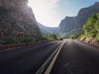 a mountain is seen behind the empty road in the mountains between trees and rocks with sun shining down on the mountain tops