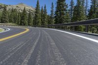 the curved road passes a mountain with pine trees and hills in the background at the base of the cliff