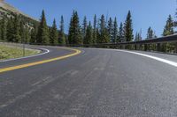 the curved road passes a mountain with pine trees and hills in the background at the base of the cliff