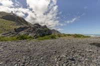 a beach covered with rocks and vegetation near mountains with clouds above it that is surrounded by lush green shrubs