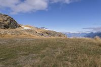 an open grassy area next to a rocky cliff face and a forest below with snow on the ground