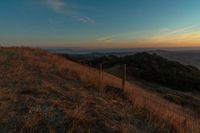 the sun rising over the mountains above the grass covered hills behind a fenced in area