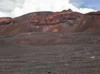 a person with an orange backpack walking on a hill in the mountains with red rocks and grass and dirt and red soil and a white cloud